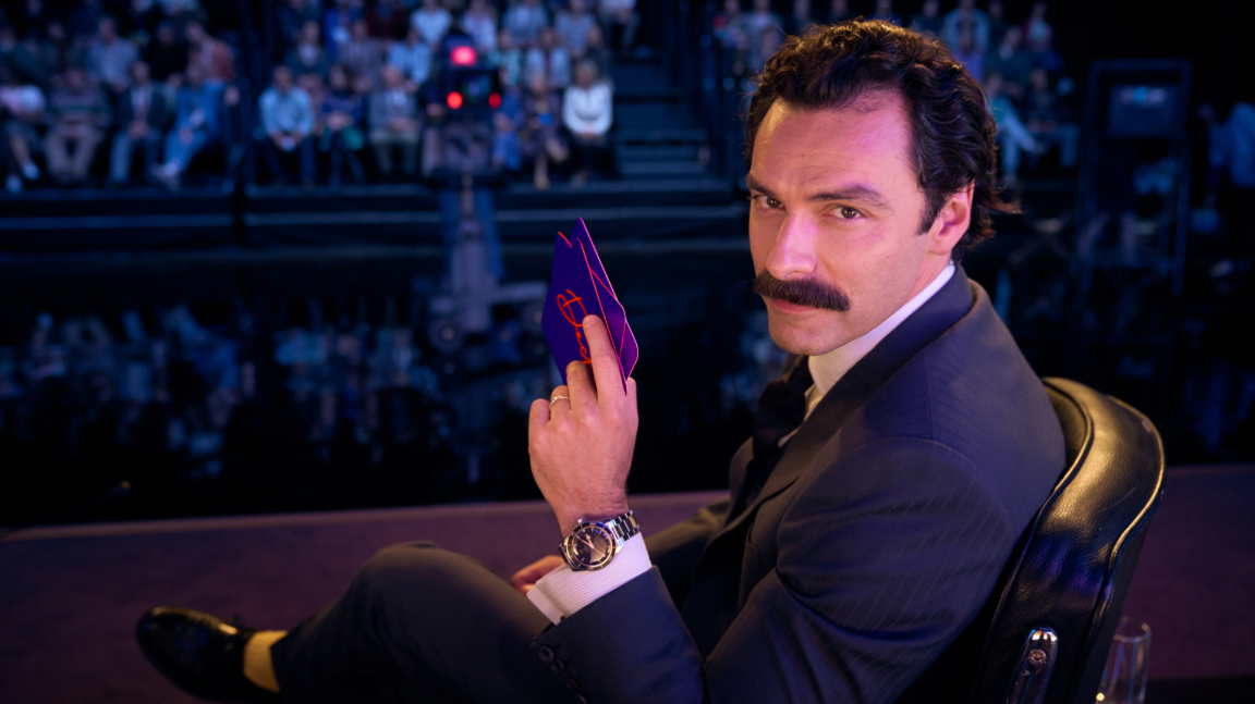 A publicity shot of Aidan Turner on the set of a 1980s TV studio in a presenter's chair with some cue cards in his hand, looking over his shoulder with a suave smile