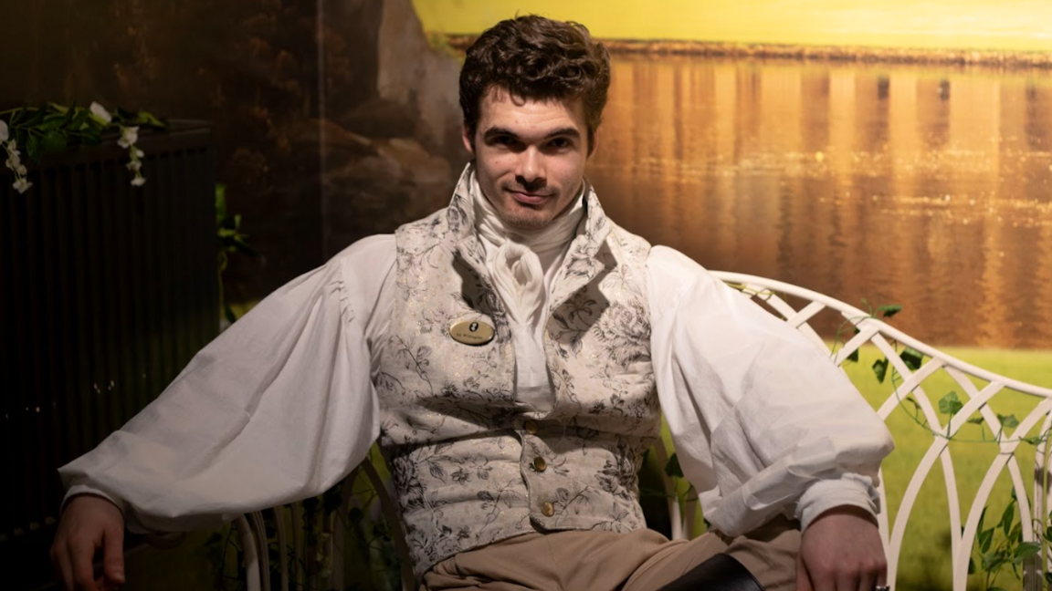 A man dressed as Mr Darcy in 18th Century clothes, reclining on an iron garden bench with a painted background of a lake behind him  