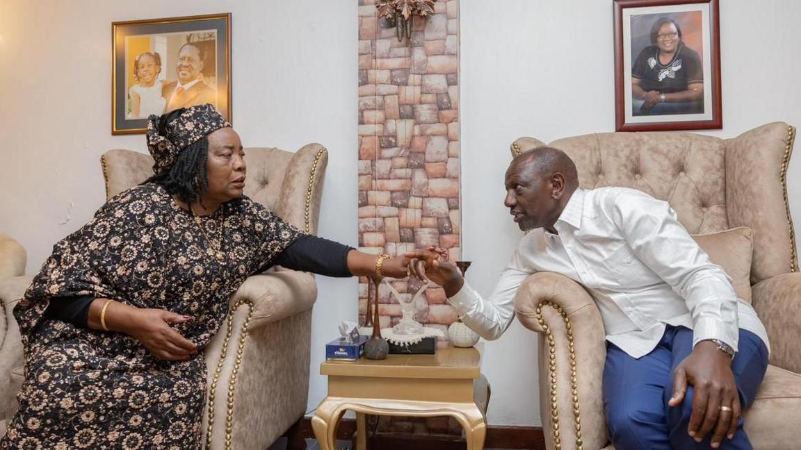 Raila Odinga's widow, Ida Odinga (left) in a patterned dress speaks with Kenyan President William Ruto (right) in a white shirt and blue trousers. Both are seated on a sofa next to each other