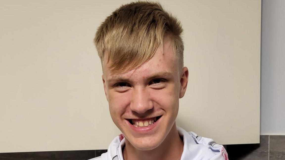 A teenage boy with light blonde hair and wearing a white t-shirt smiles at the camera.