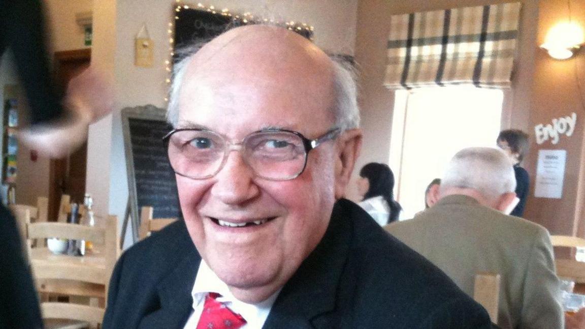 Donald Dixon smiling at the camera. He has receding white hair, glasses, and is wearing a black suit jacket, a white shirt and red tie. 