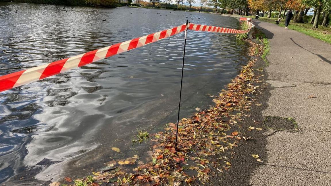 Pontefract Park lake, with leaves washing up on the side, a red and white safety barrier, and a woman walking in the distance