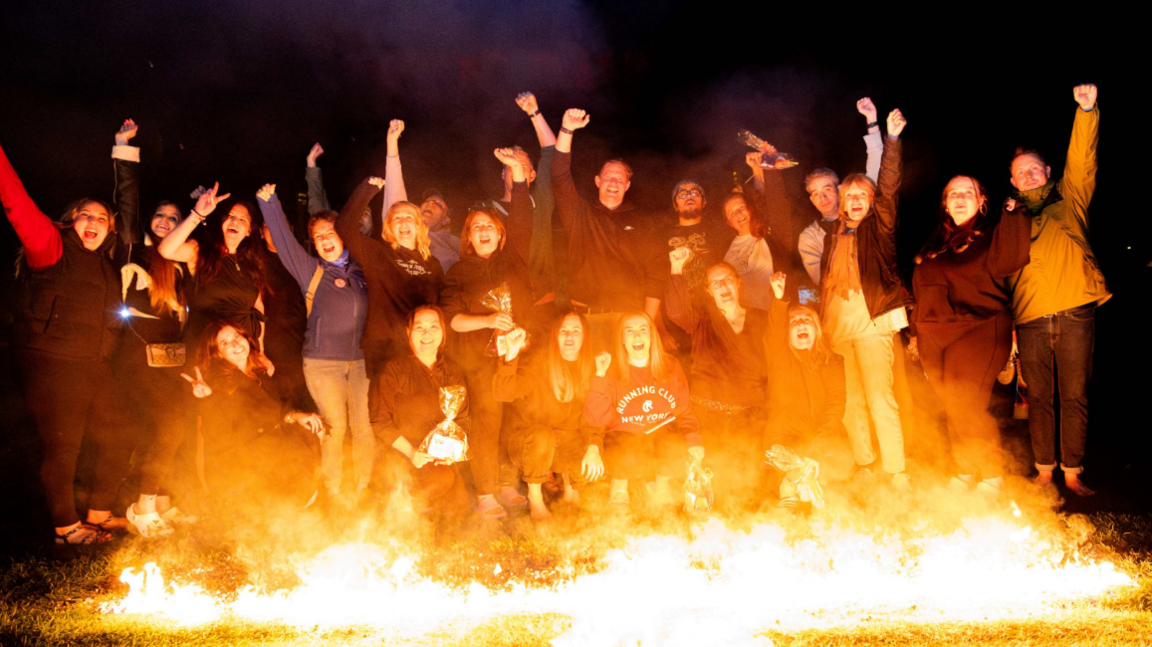 About 30 people stand in front of a line of coals which are on fire and raise their hands in the air in jubilation. It is dark and the sky is black behind them. Their faces are lit by the flames.