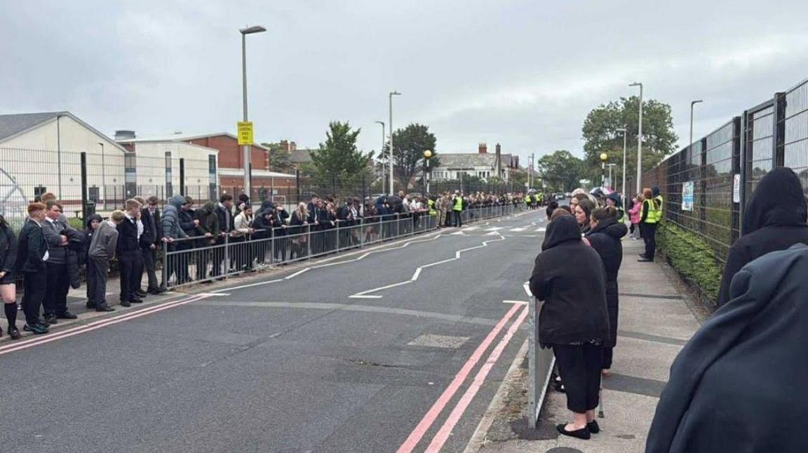 Hundreds of school pupils wearing uniform line the streets surrounding Armfield Academy in Blackpool. They are paying their respects ahead of the funeral cortege for teacher Pat Burns.