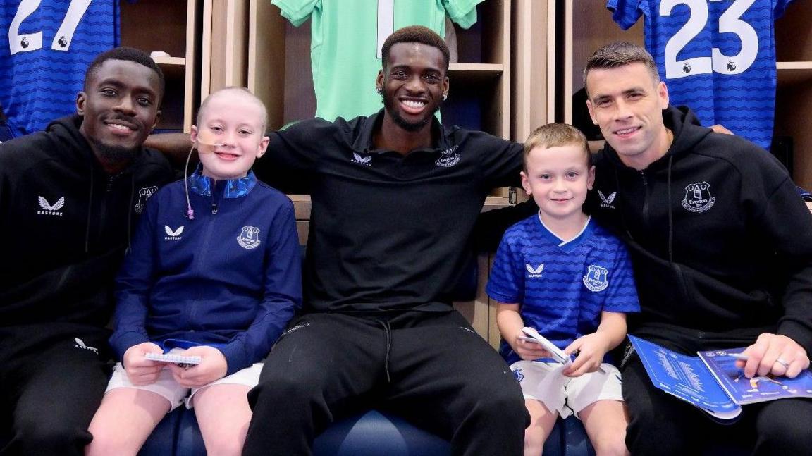 Mason and his younger brother are both wearing Everton FC strips. They are sitting in the team's dressing rooms next to Idrissa Gana Gueye, Tim Iroebugnam and the club's captain Seamus Coleman