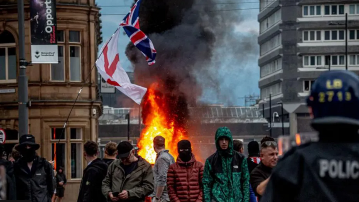 Crowds gather in front of a burning building, with riot police visible in the corner of the image