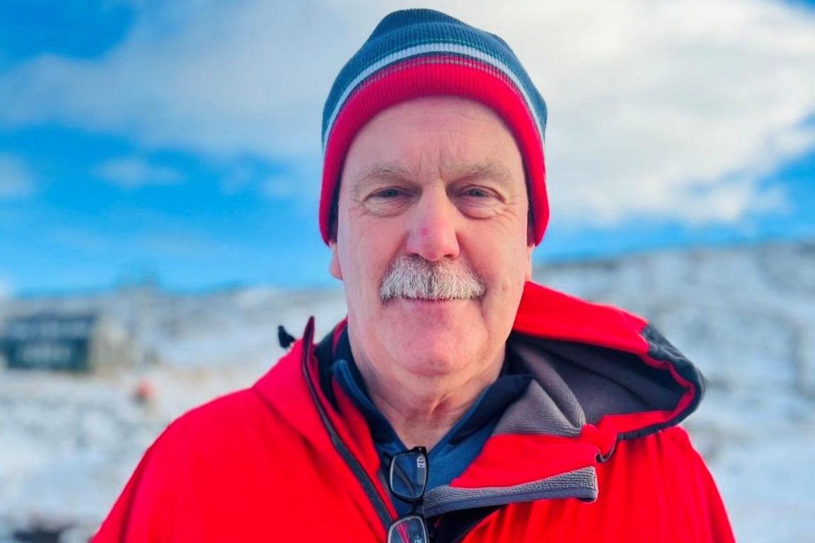 Man with grey moustache looking at camera, wearing red, white and blue warm wool hat, and a bright red jacket, with a snowy scene behind and a cloudy blue sky.