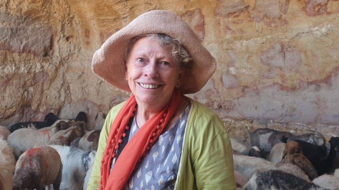 A woman smiling at the camera wearing a hat, red scarf, green jumper and a blue top. You can see a group of sheep behind her and she appears to be in a cave.