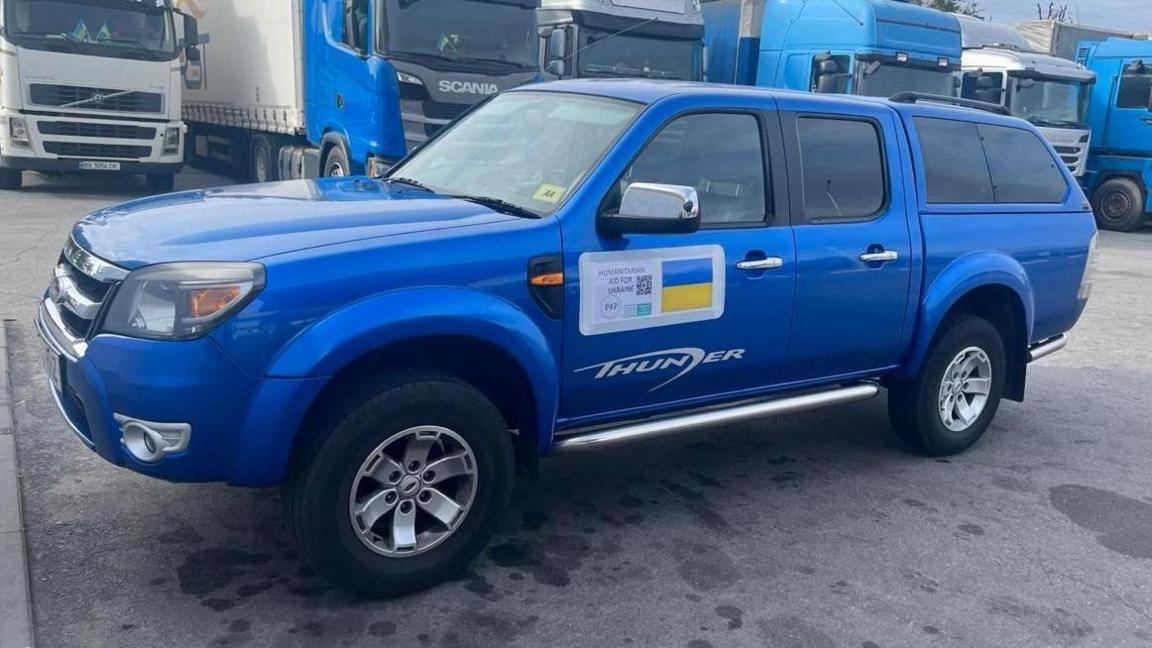 A bright royal blue metallic Ford Ranger pickup-style car sits in a big car park, possibly at a port, with trucks lined up in a row behind it. It has an official sticker on the side with a Ukraine flag and a bar code to identify the car