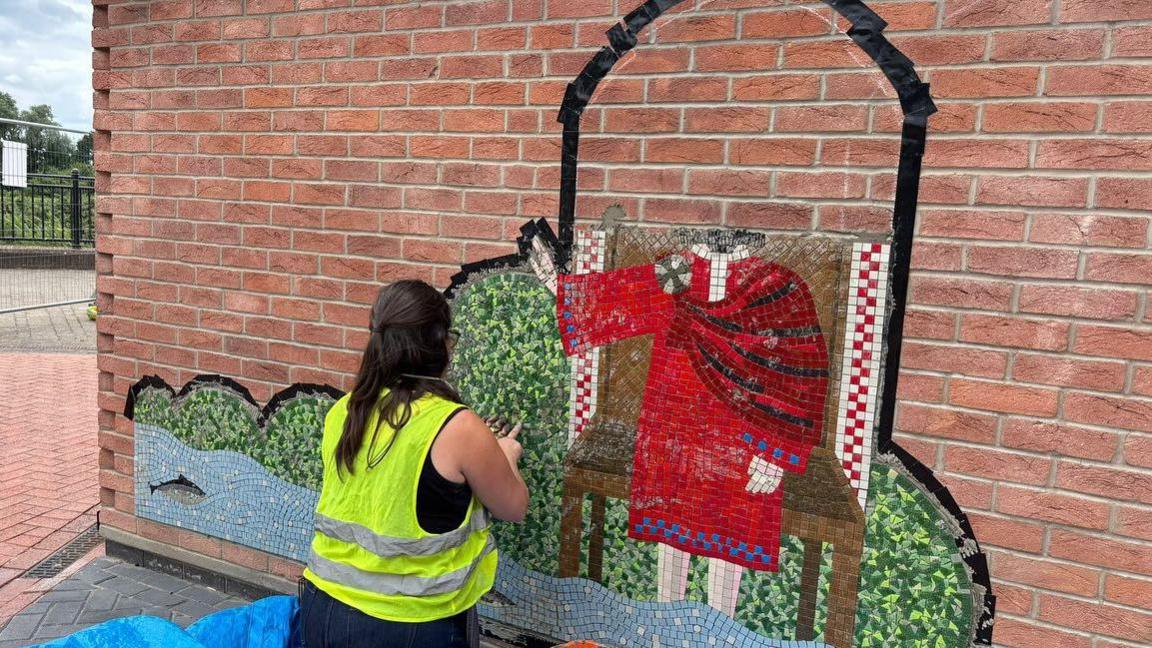 A woman with long brown hair and wearing a yellow hi-vis vest has her back to the camera. She is working on the part-completed mosaic.