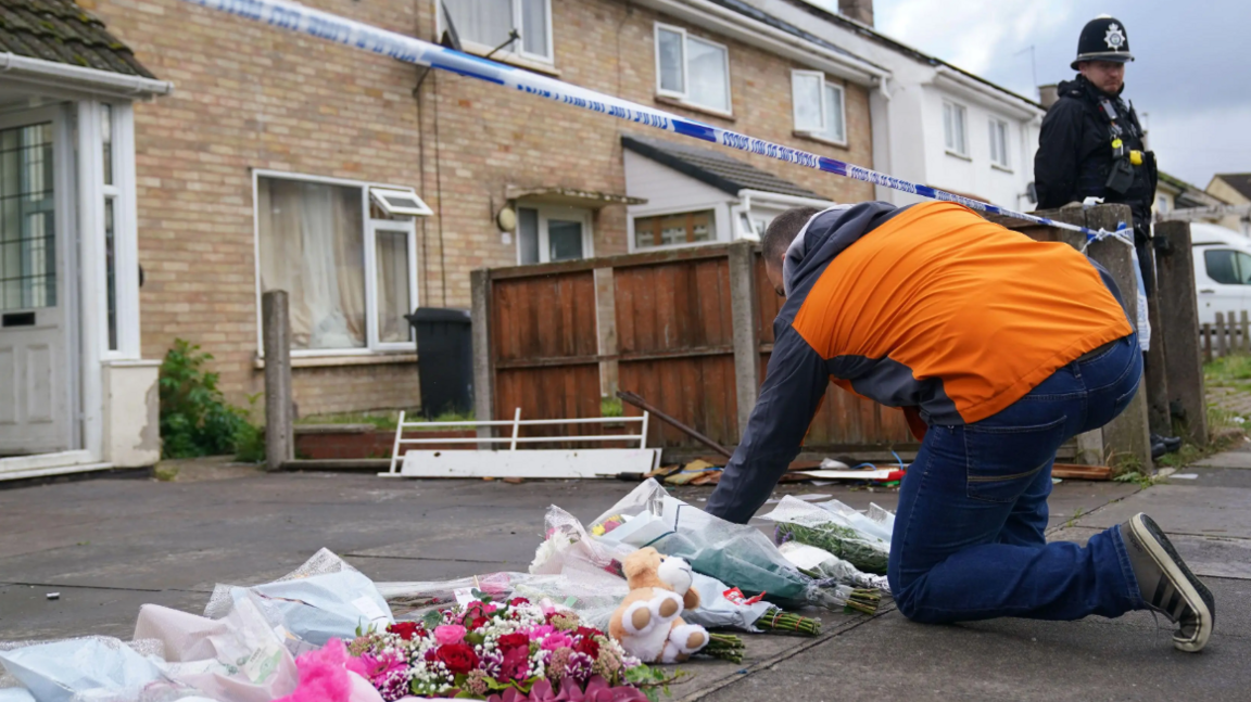 A man in an orange coat and jeans placing flowers on paving slabs outside a light brick house with police tape outside and a police officer in full black uniform stood near by