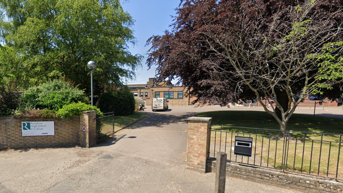 Front of school gates showing white sign which reads Reepham High School & College, a driveway, trees and a parked white van