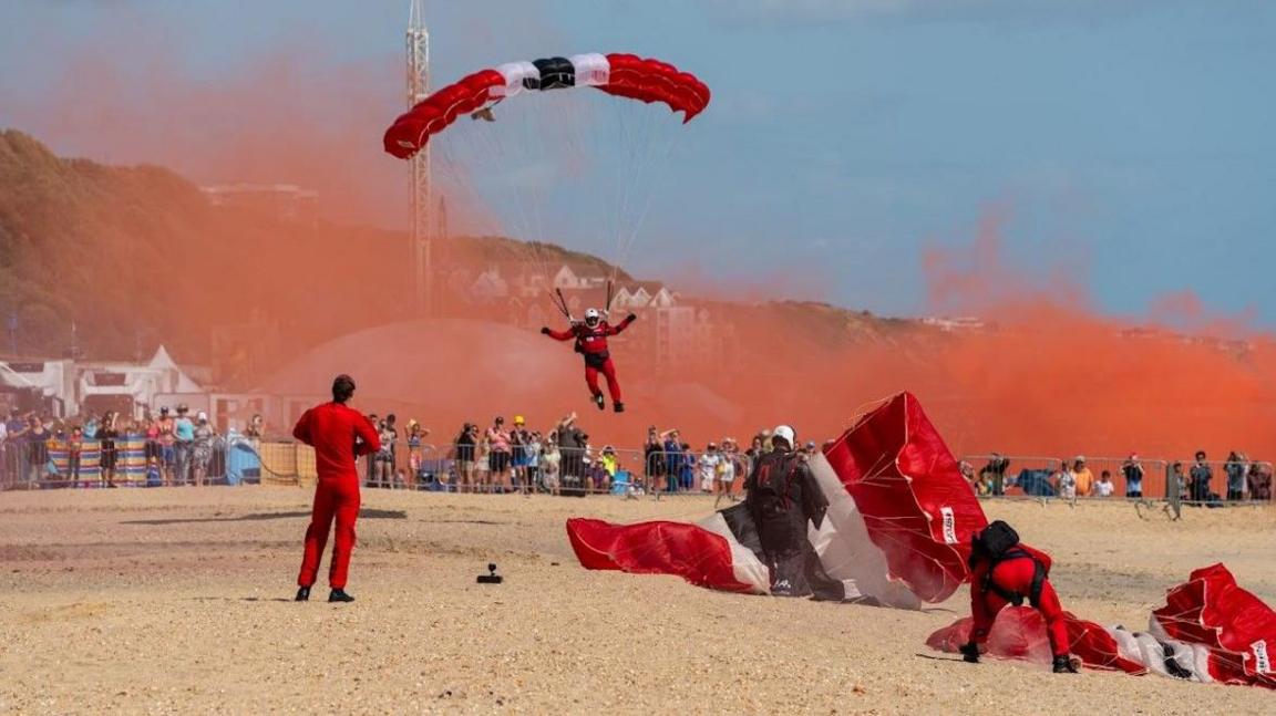 Red Devils parachutists landing on Bournemouth beach with spectators watching from behind barriers. The parachutists are wearing read overalls and have red and white parachutes and are trailing red smoke behind them