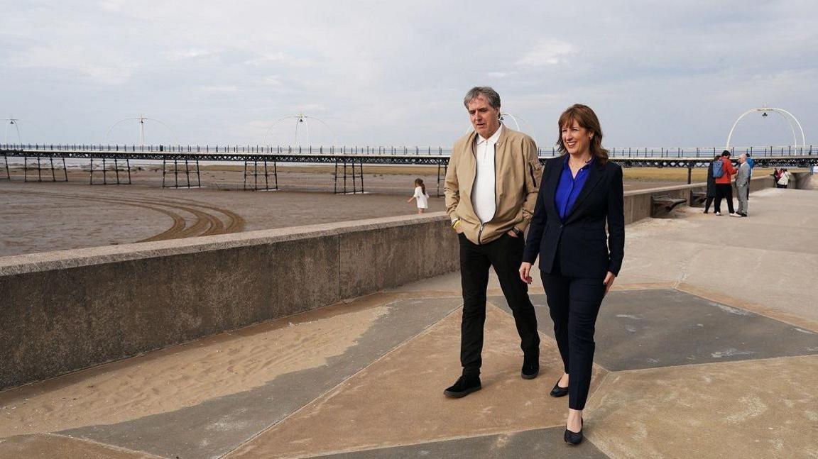 Liverpool City Region mayor Steve Rotheram and Chancellor Rachel Reeves stroll on the promenade with the pier stretching out behind above a sandy beach. Some people are milling around in the background, where a child also plays and runs.