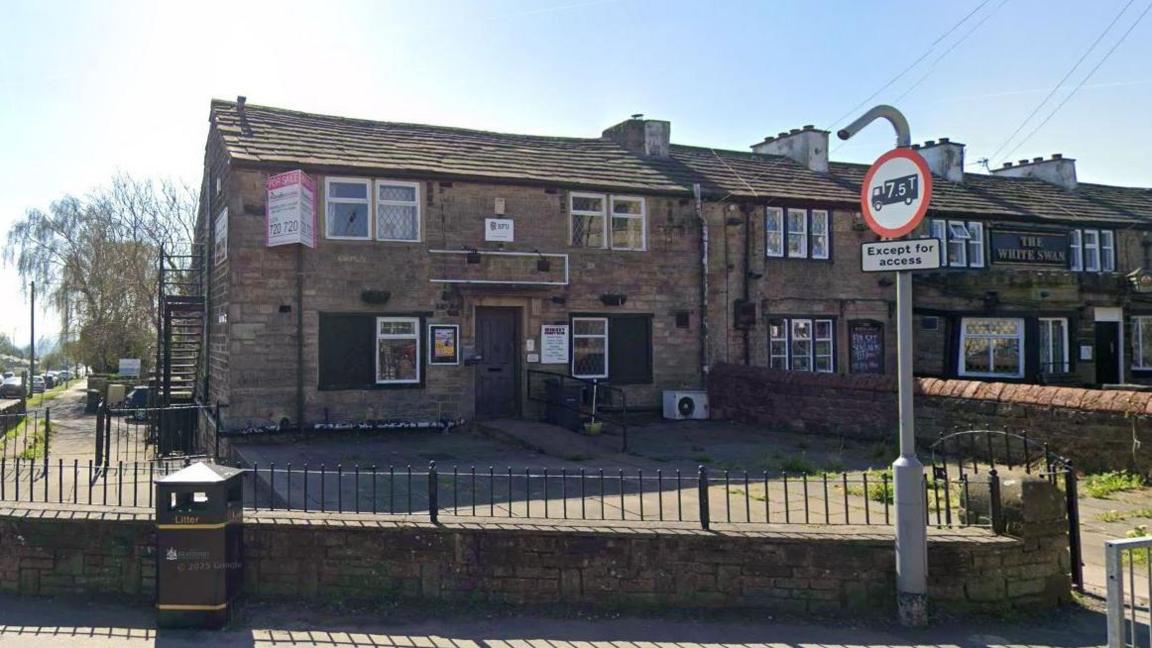 A street corner with a two-storey stone building with multiple sash windows and signs. Posters and notices are visible on the exterior walls. It is attached to a similar building that has a sign saying The White Swan on the front. In front of the building is a fence, a black bin and a road sign.