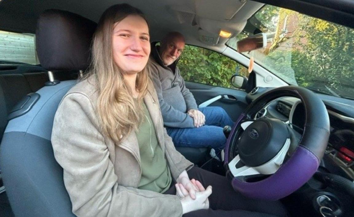 Zoe Johnston on the left and Carl Goodwin in a car, with Ms Johnston smiling, as she sits in the driver's seat, with a black and purple steering wheel in front of her.