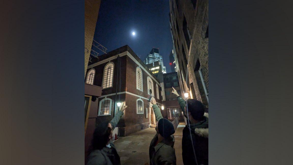 Three men stand outside the synagogue at night and point towards the moon, which appears full.