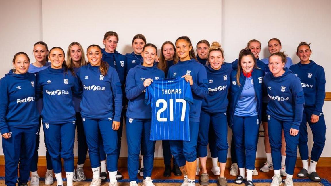 The Ipswich Town Women's team are pictured standing together in a group. Natasha Thomas is stood in the middle holding her anniversary kit. The top has her name on the back as well as the number 217 marking her number of appearances. 