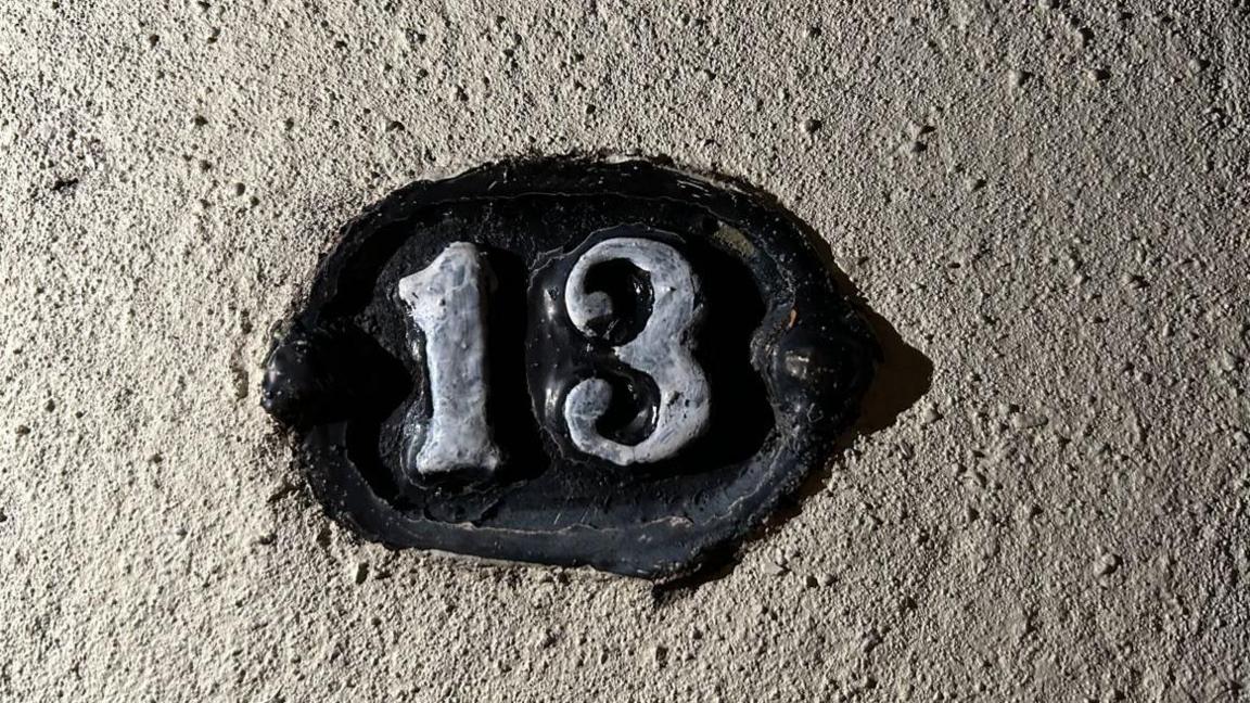 A close-up of a number 13 sign of a house. The number is painted in grey with a black circle around it. The wall is white and bobbled