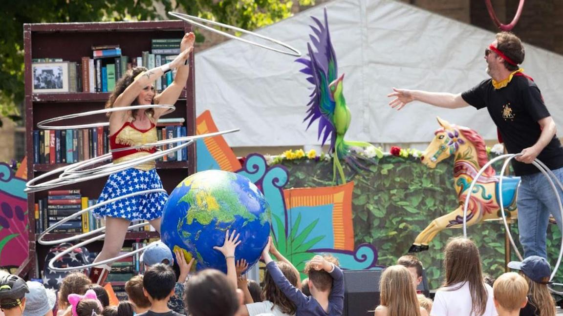 Two acrobatic performers putting on a show in front of a crowd of children. The man on the right, wearing blue jeans and a black t-shirt, is throwing silver hula hoops to a woman on the left, who is wearing a wonder woman outfit. The children are throwing around an inflatable globe, and in the background there are carousel horses, a large parrot, and a wooden shelf stacked with books.