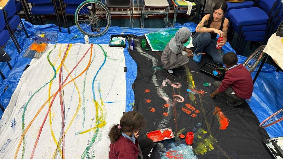 Children and a workshop leader work with different paint colours and bicyle wheels to create a work involving bicycle tracks on the floor.