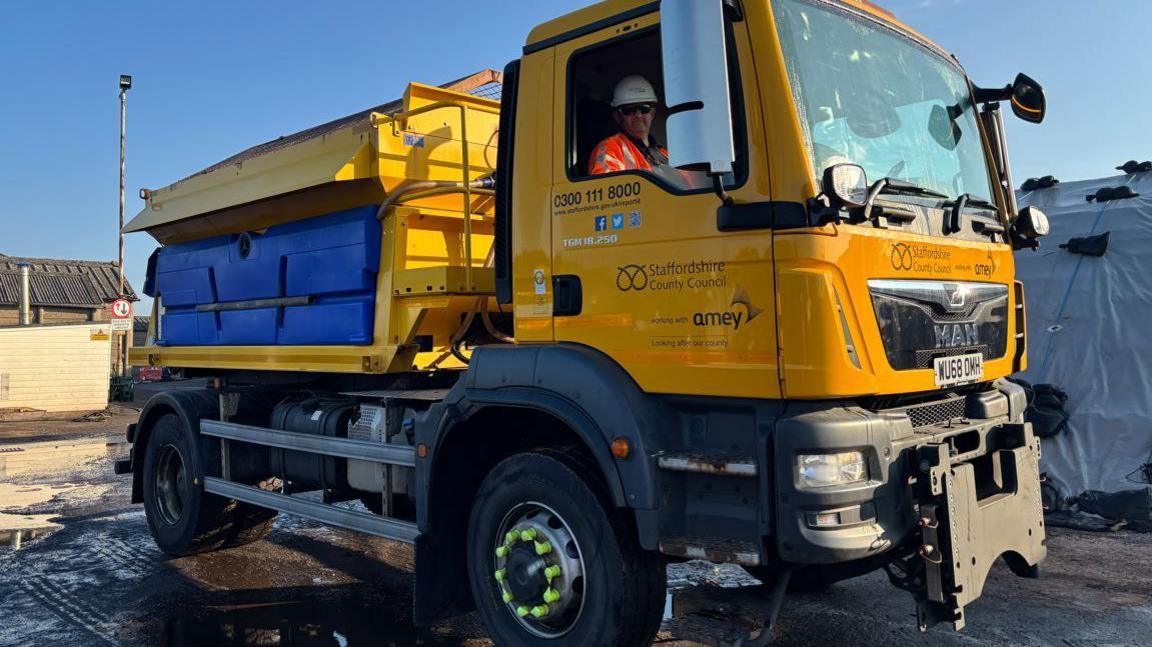 A yellow gritting lorry with Staffordshire County Council written on the side. A male driver in an orange high-vis jacket and white hard hat is sitting in the front. The sky is blue in the background and the lorry is parked in a yard.