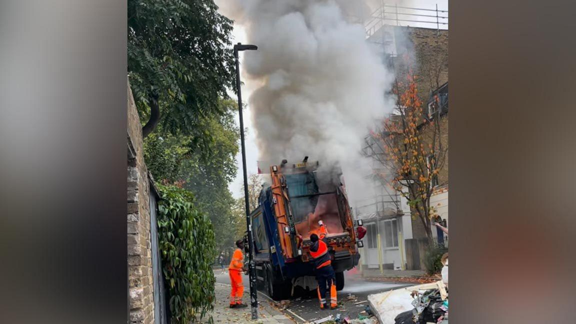 Refuse workers use hand held fire extinguishers to try and put the fire out in the back of a bin lorry. A large amount of smoke and some flames can be seen