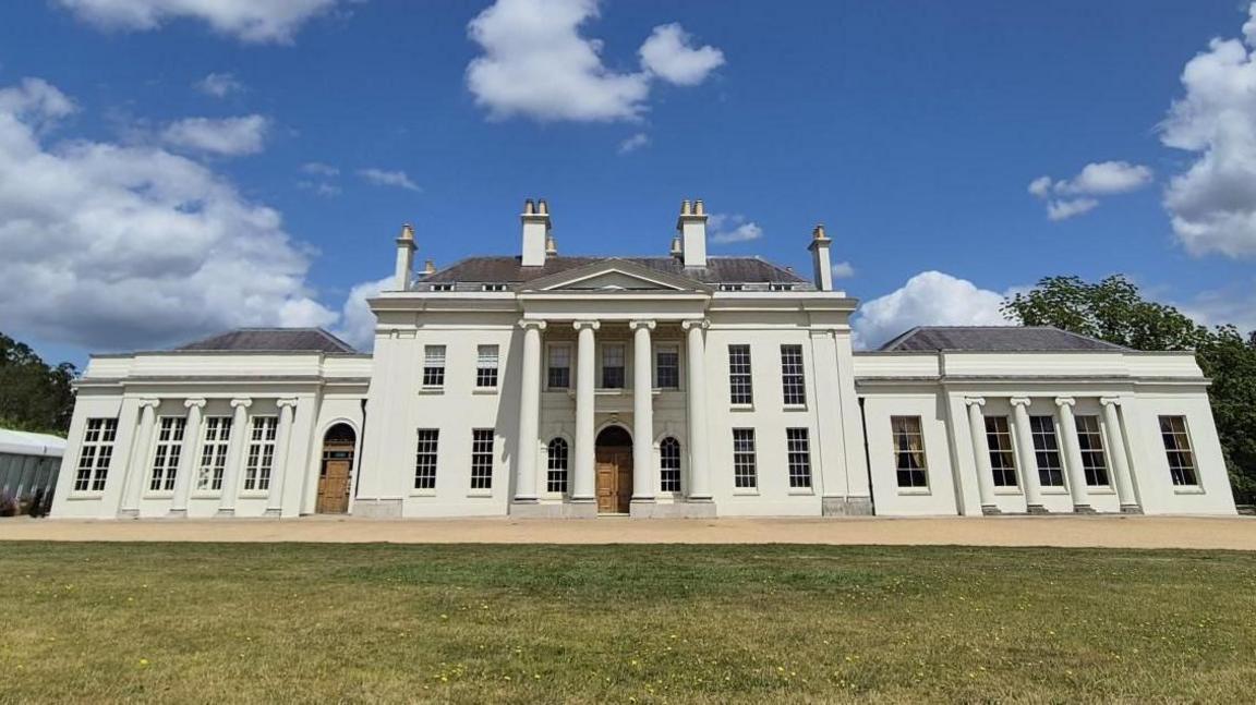 A large white building with columns has a grey roof and 4 chimney breasts on top of it. A lawn is in front and there is a blue sky above with some clouds and trees to the side.