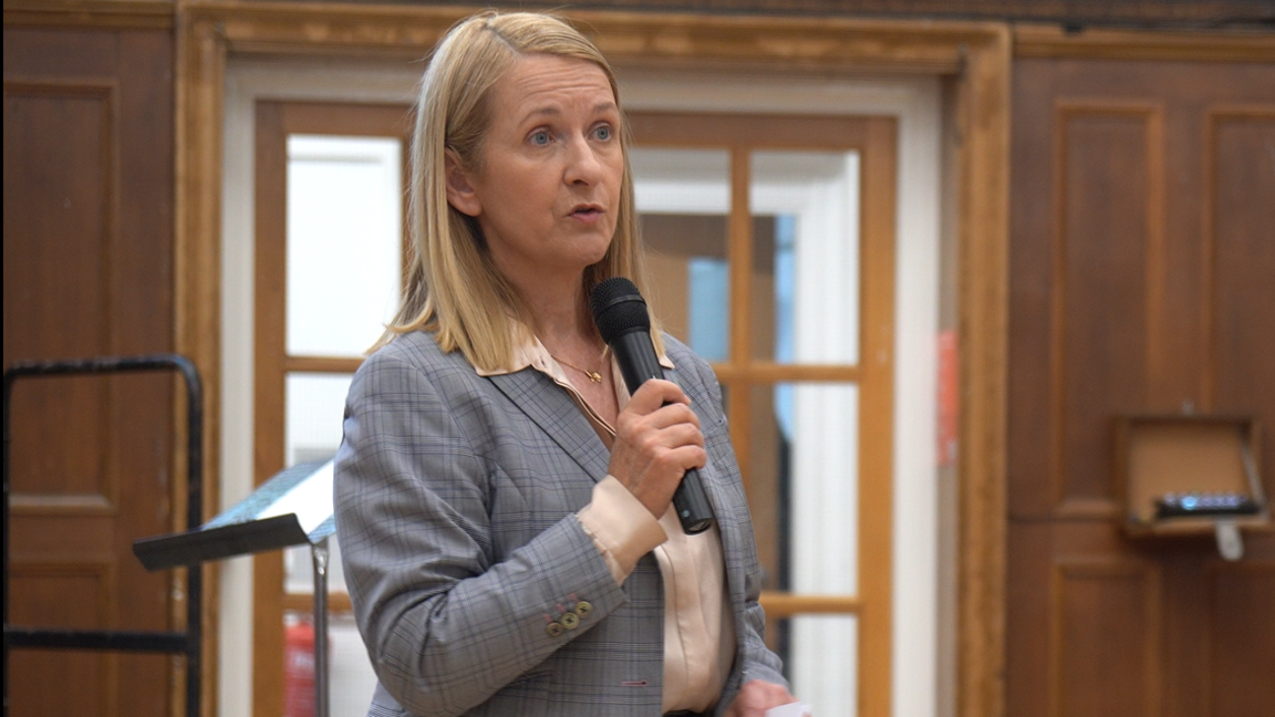 There is a woman with blonde shoulder-length speaking into a microphone, wearing a plaid grey blazer and light pink shirt. She is also in a hall, with wooden walls behind her and what appears to be glass windows.