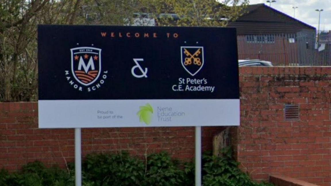 School sign at entrance for Manor School and St Peter's Academy. Both have logos on the sighn, which has a blue background and a white band at the bottom. A school building is visible behind.