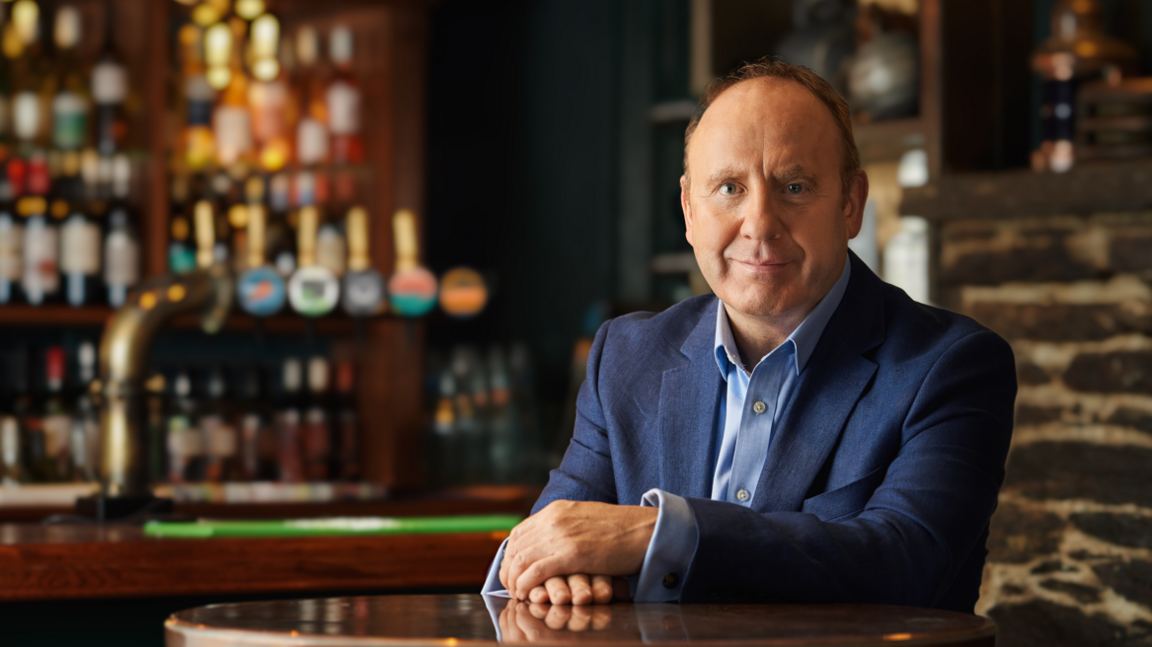 Kevin Georgel - wearing a blue suit sits at a table inside a pub. The bar is out of focus over his right shoulder.