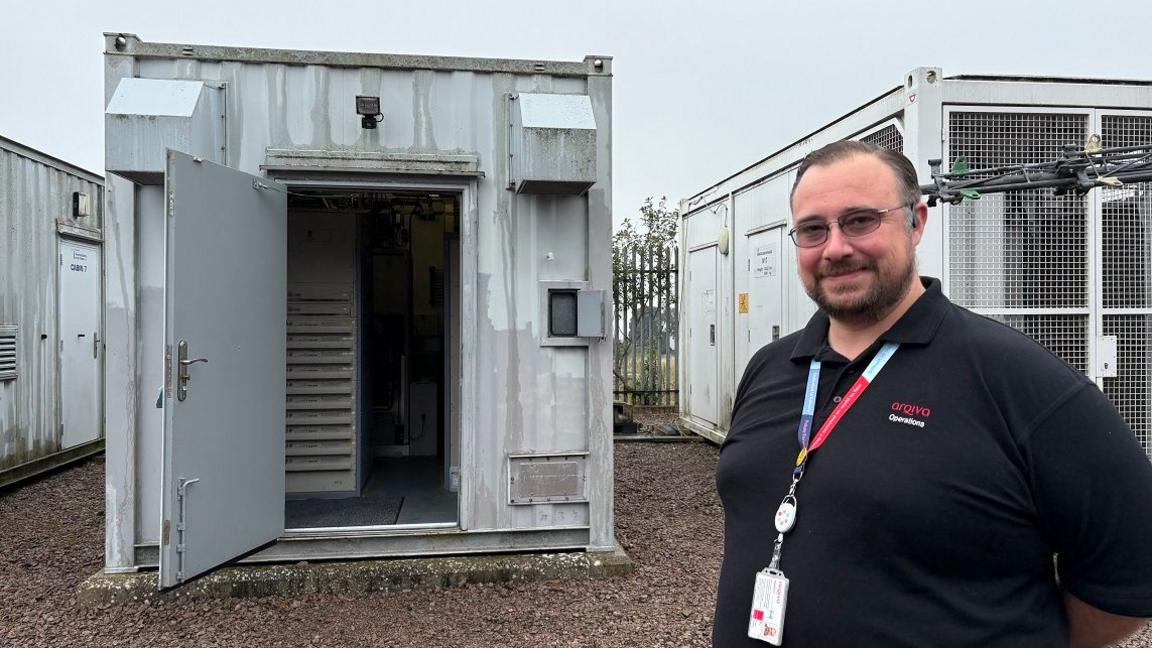 Alan Storer with short dark hair and beard wearing a black Arqiva T-shirt and a red lanyard standing outside a square grey metal cabin with its door open. An equipment rack can be seen inside. There are more grey cabins on either side, which can be seen to extend some distance from the door.