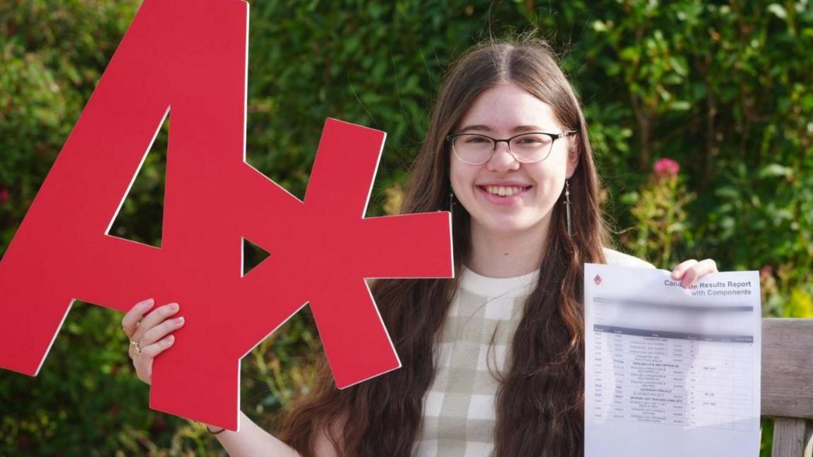 Gabrielle Morgan has long dark hair and glasses. She is holding up a big red A* sign in cardboard and holding the letter detailing her results. She is smiling and there is a garden behind her. 