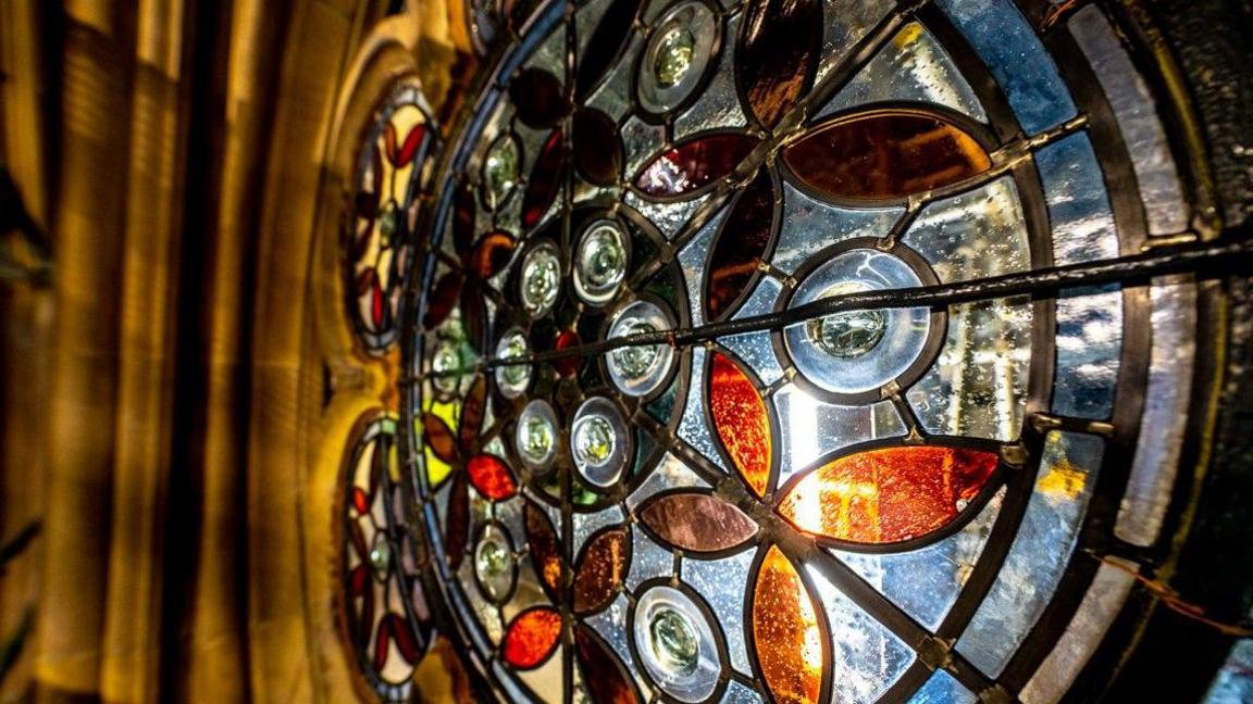 A close up of a stained glass circular window which includes amber and brown coloured leaves.