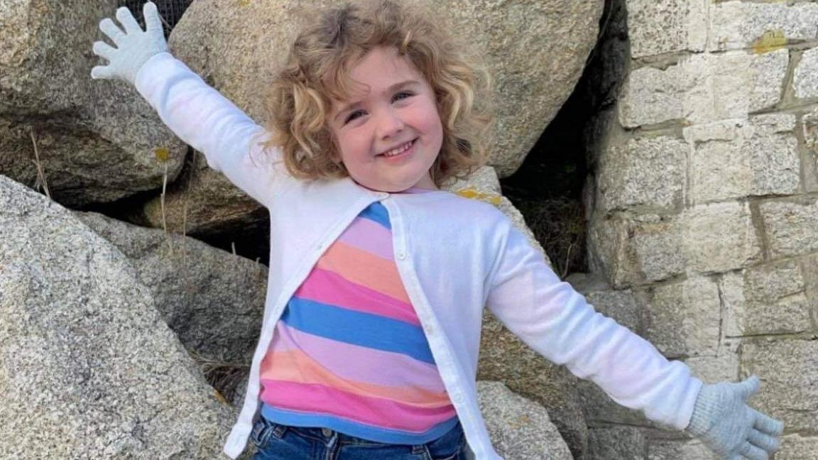 Amber is standing on a big rock surrounded by other large rocks on the beach. She is holding her hands out high away from her body like a star. She is grinning as she poses with one of her legs slightly bent for emphasis. She has curly blonde shoulder-length hair and blue eyes, She is wearing a denim cut-off skirt over pink tights, blue trainers and a pink cardigan over a brightly coloured stripy top.