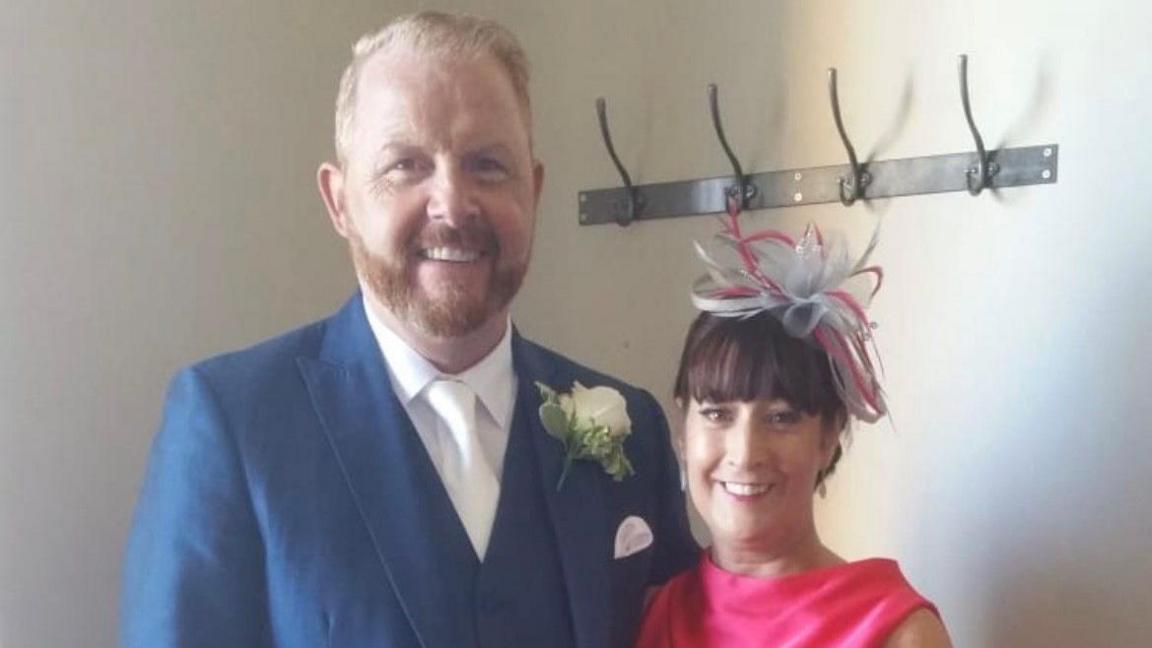 A husband and wife on their wedding day. The man, on the left, is of stocky build, has fair hair and is wearing a blue suit. His wife, on the right, is wearing a feathered hat and a pink dress. They are standing against the backdrop of a coat hanger.