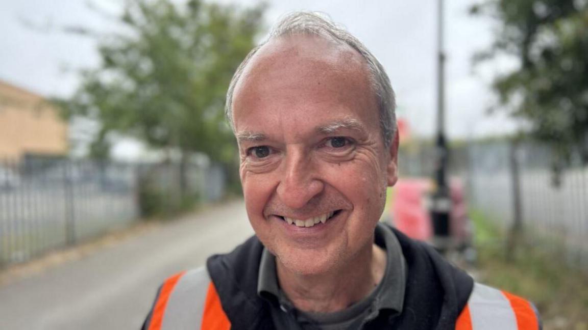A man with short grey hair is smiling into the camera for a head and shoulders shot. He is wearing orange high vis, a black jumper and polo shirt. Behind him is an alleyway with blurred metal fences on both sides.