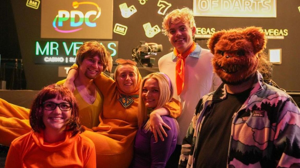 Six people are in the photo, including someone on the right with a bear face mask on. The woman who is the third person from the left has her arms around the two people either side of her.