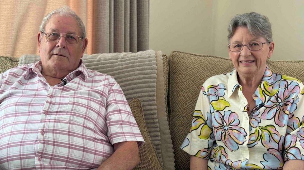 A man in a white and red checked shirt is on the left next to a woman with a yellow shirt that includes large images of flowers. They both have glasses and grey hair and are sitting down looking at the camera.