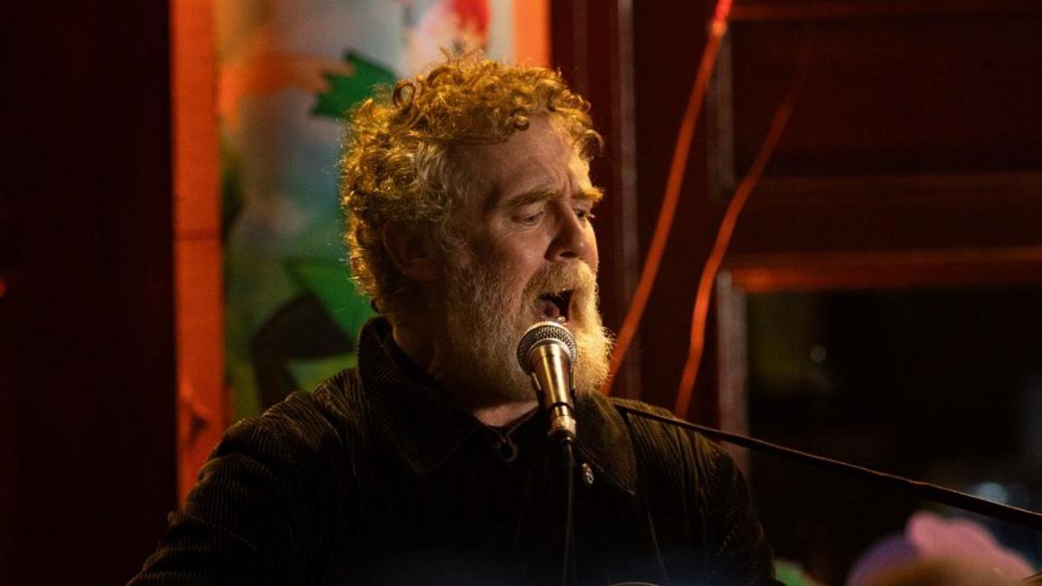Glen Hansard has curly reddish/grey hair and beard and is singing while playing a guitar