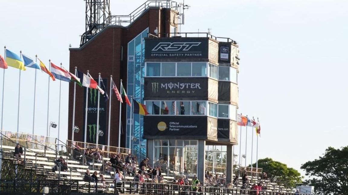The grandstand, which has a handful of people sitting in it. There is tower in the middle with large windows around it, which have sponsorship banner beneath them. There is also a row of international flags along the back of the seating area.