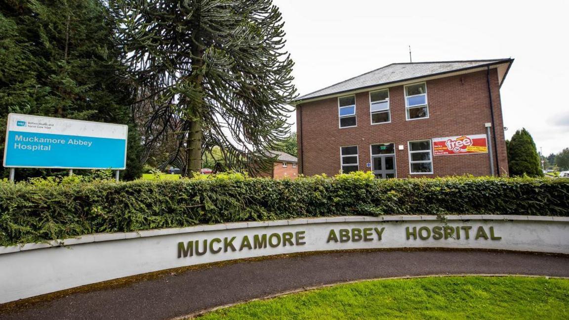 A white wall is pictured in front of the hospital with writing saying "Muckamore Abbey Hospital".  There is a blue and white sign to the left with the hospital name on it also. In the background there is a red brick building and to the left of it trees.