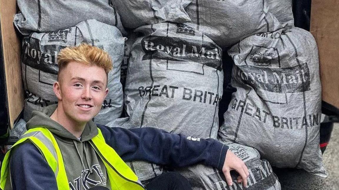 Jamie is sitting in front of grey Royal Mail bags that are in the back of a van. He is wearing a green and navy hoodie with a high visibility jacket. The bags are all filled and are stacked three wide within the vehicle. One bag is on the ground. 