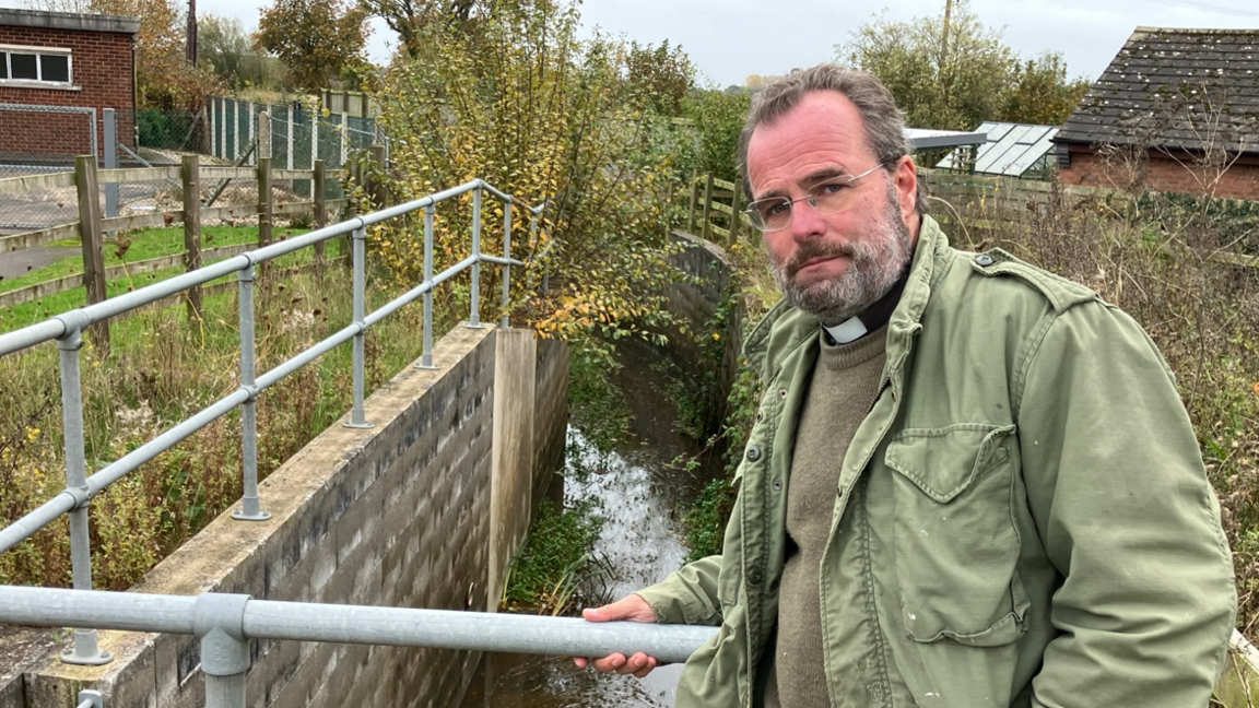 A man with a beard and glasses wearing a green jacket and a vicar's collar standing in front of a culvert with a brick wall and metal railings surrounding it