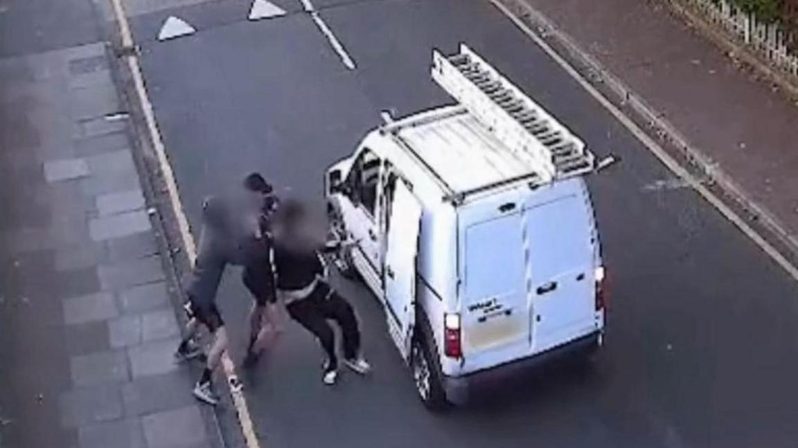 An ariel CCTV view of Havelock Road in Luton. A small white van with a ladder on the roof is stopped in the middle of the road, with a side door panel open. Next to the door, two men try to drag a third into the van. Their faces are all blurred out and you can see three pairs of feet. The victim wears black jogging bottoms with white stripes.