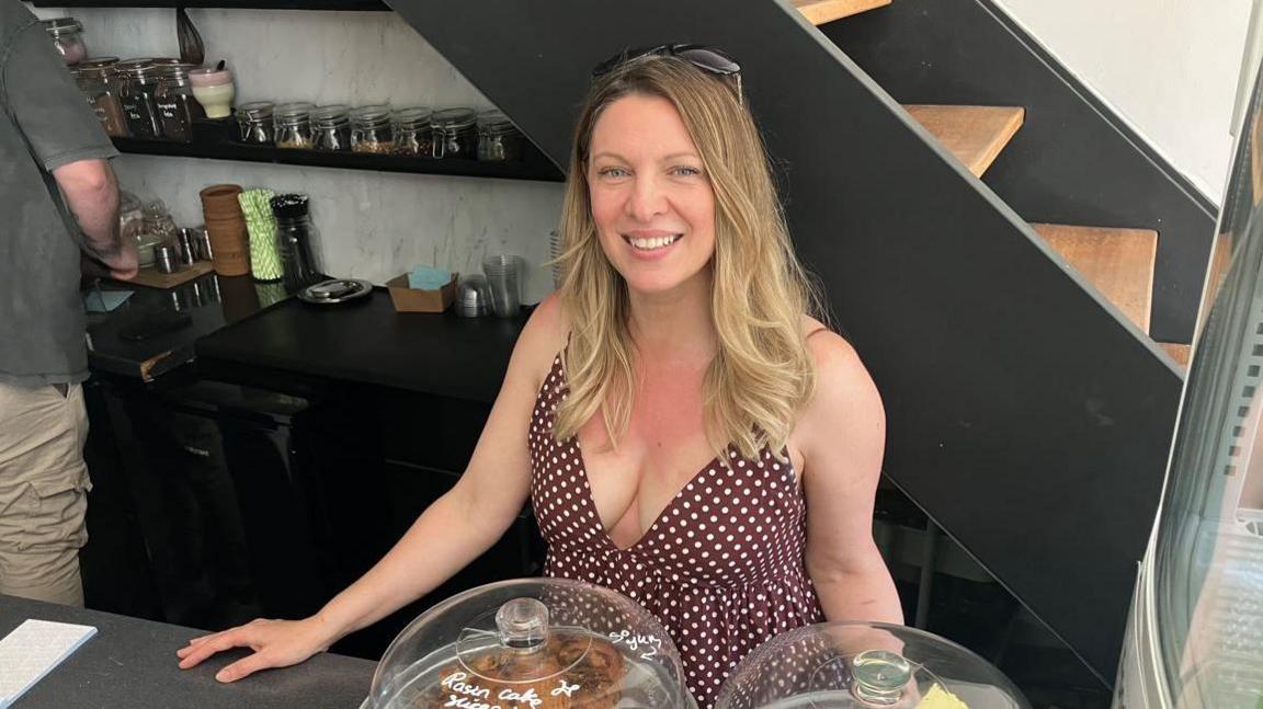A woman with blonde hair and a maroon dress with white spots is stood behind a cafe counter with cakes under class domes. Behind her are shelves with jars on, a person making a drink and some stairs.