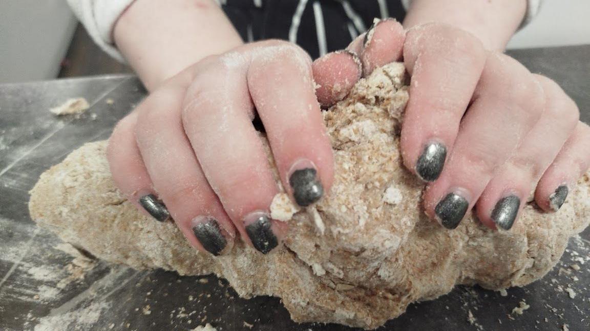 A woman with black painted fingernails kneads some pizza dough.