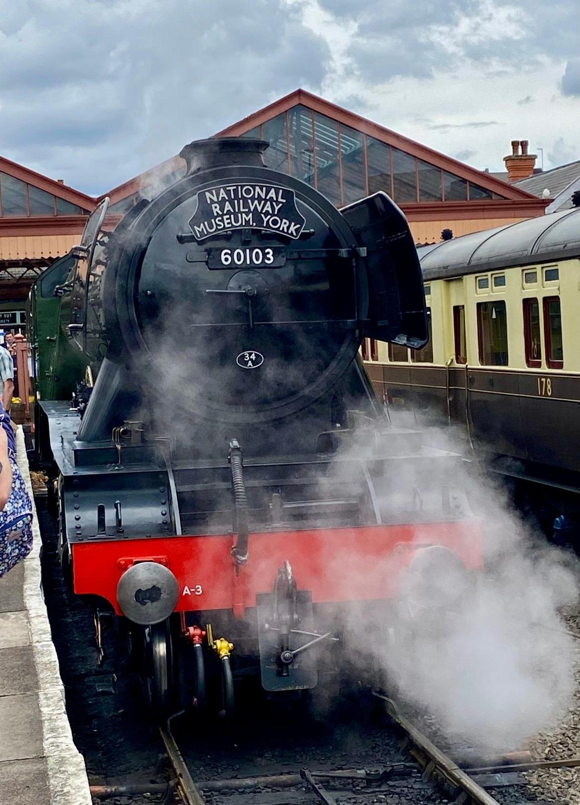 A steam engine with a black front and red buffer panel sits in a station next to a carriage with steam coming from the engine. On the front is a sign with "National Railway Museum, York" on it.