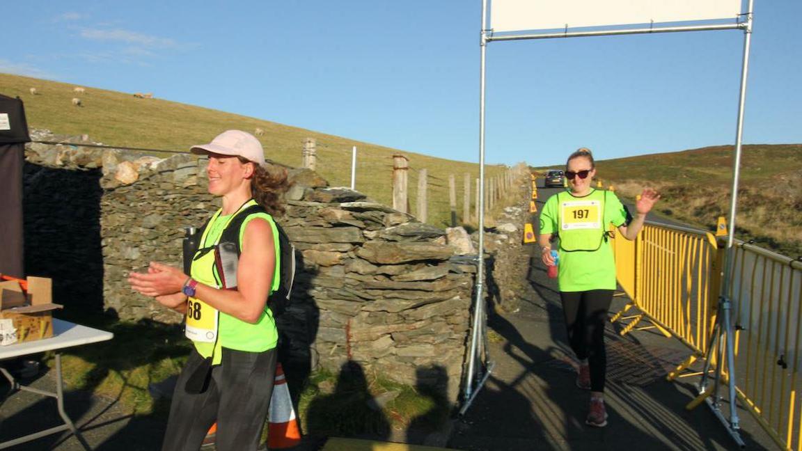 Two women are walking, they both wear high vis vests, you can see a barrier on the road, they both smile as they reach the finish line.