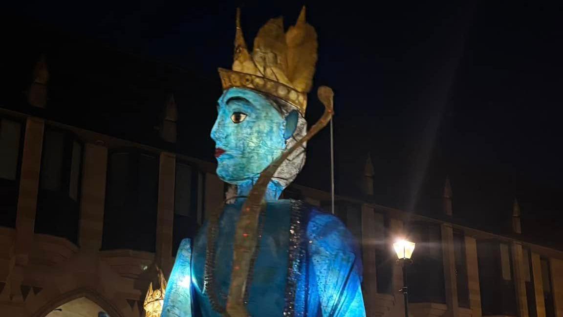 A large scale blue, and gold puppet, light up, in the centre of Northampton, with a street light behind it, and a large building.
